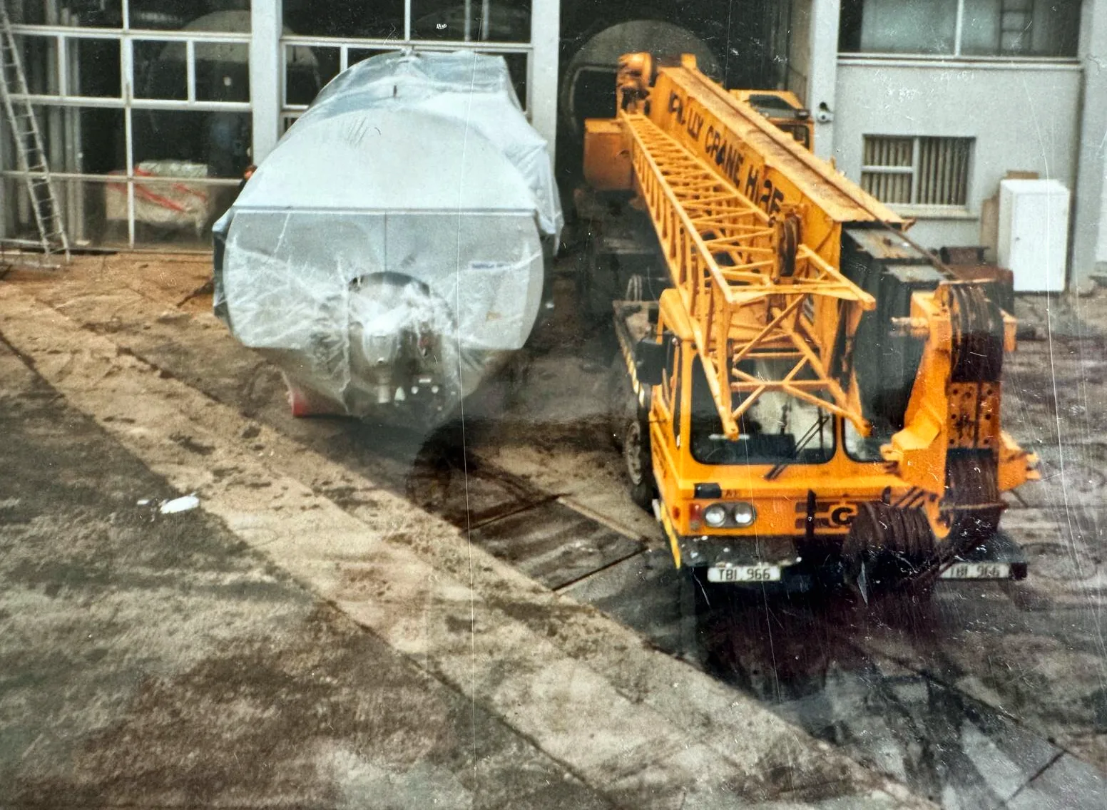 Archive photograph of a McNally’s Crane Hire truck crane beside a plastic-wrapped steel vessel outside an industrial unit