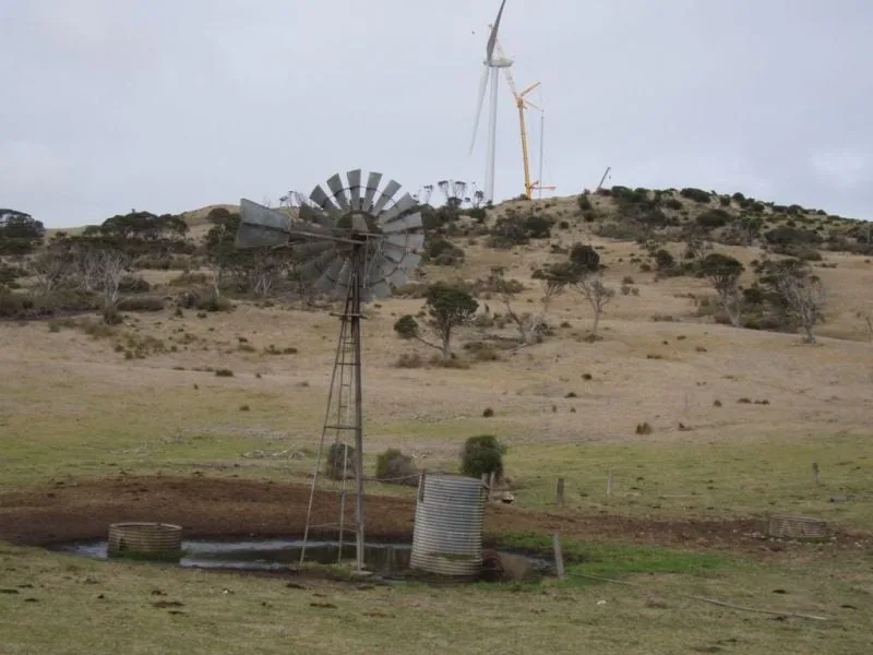 An old farm windpump and water tank in the foreground, with a crane erecting a wind turbine on the hill behind